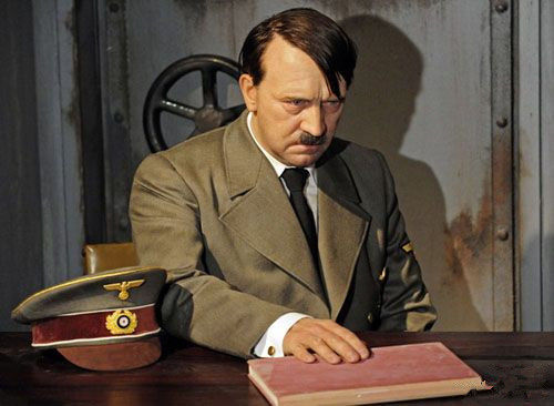 蜡像希特勒身处幽暗地堡房间，背对门坐在木质书桌前 