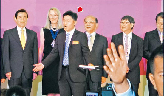 男子(箭头处)趁马英九与台中市长胡志强等人合影时冲上主席台用英文抗议,台下随扈(右前)没听懂陈说内容,忙着遮挡记者拍照角度。(图片来自台媒) 男子(箭头处)趁马英九与台中市长胡志强等人合影时冲上主席台用英文抗议,台下随扈(右前)没听懂陈说内容,忙着遮挡记者拍照角度。(图片来自台媒)